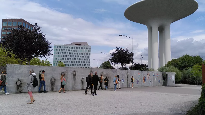 Bild tagen i Hyllie Vattenpark. På bilden är det en mur med dekorationer av mosaik och vattentornet i bakgrunden och människor utspridda som går ljudvandringen. 